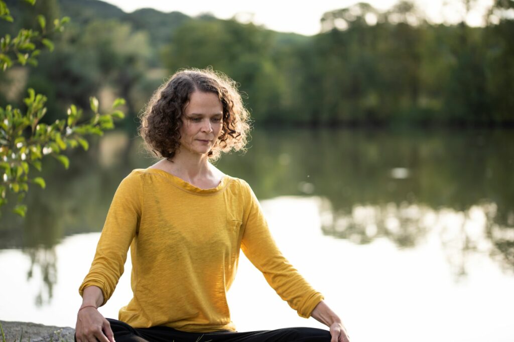 Eva Neumayer-Steiof sitzt im gelben Shirt, in Meditationshaltung in der Natur, neben dem Wasser